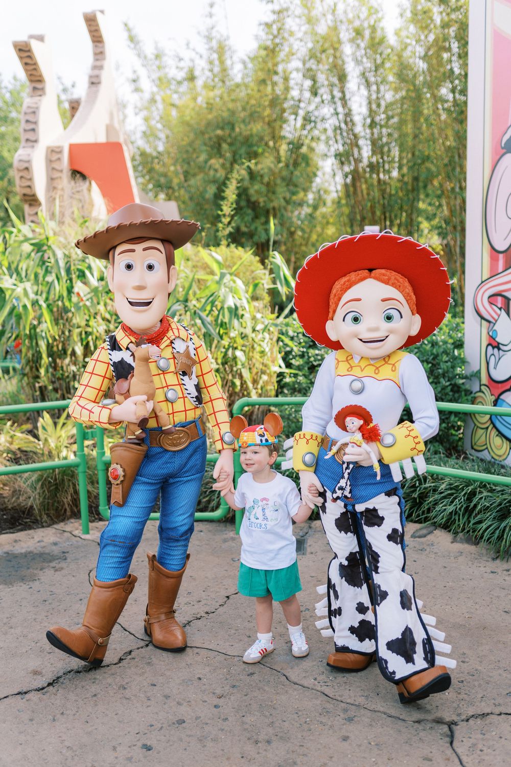 Toy Story theme park characters pose with a young visitor on a decorated pathway surrounded by greenery.