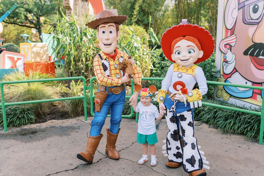 Two costumed characters from Toy Story pose with a young child at a Disney theme park with greenery in the background.
