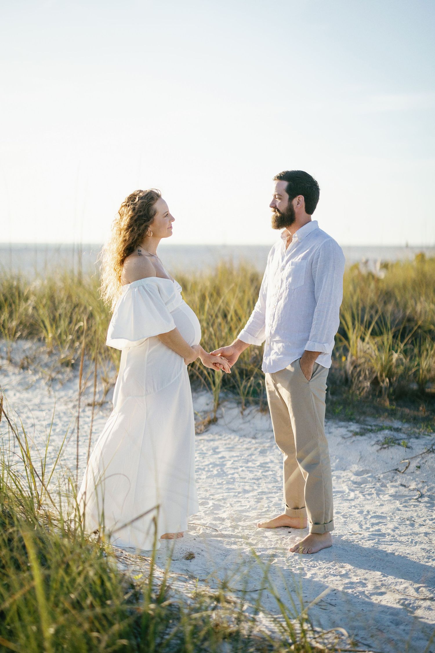 Couple in white outfits poses tenderly together on a sandy beach with coastal grasses during golden hour.