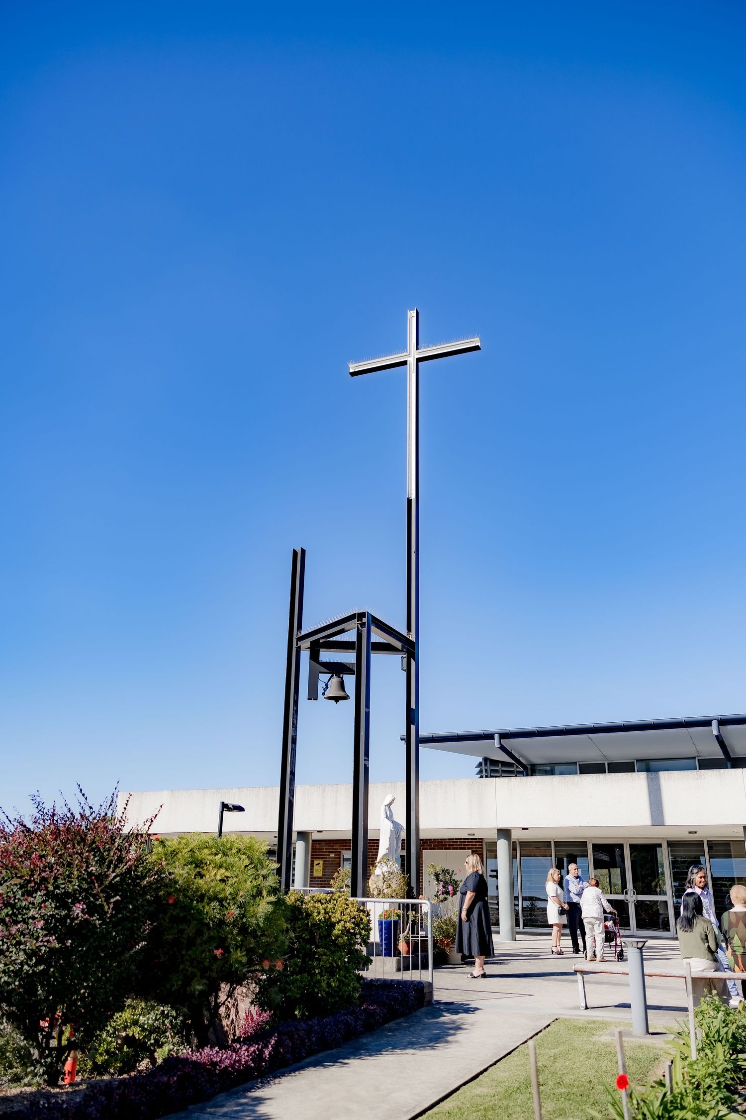 A tall white cross and bell tower stand against a bright blue sky outside a modern church entrance.