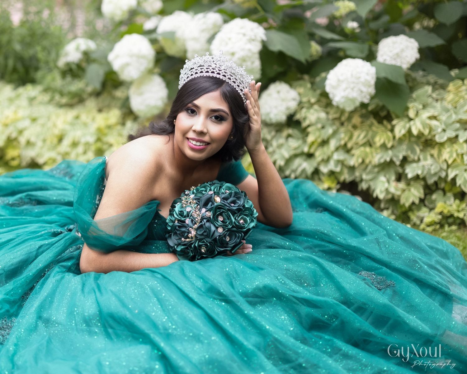 A flowing teal quinceañera ball gown with beaded embellishments photographed in a garden with white hydrangeas blooming.