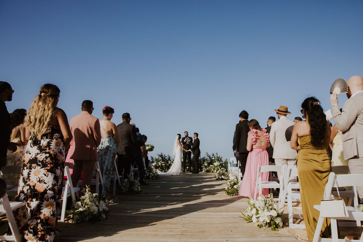 Wedding guests gathering on a boardwalk for an oceanside ceremony under blue skies.