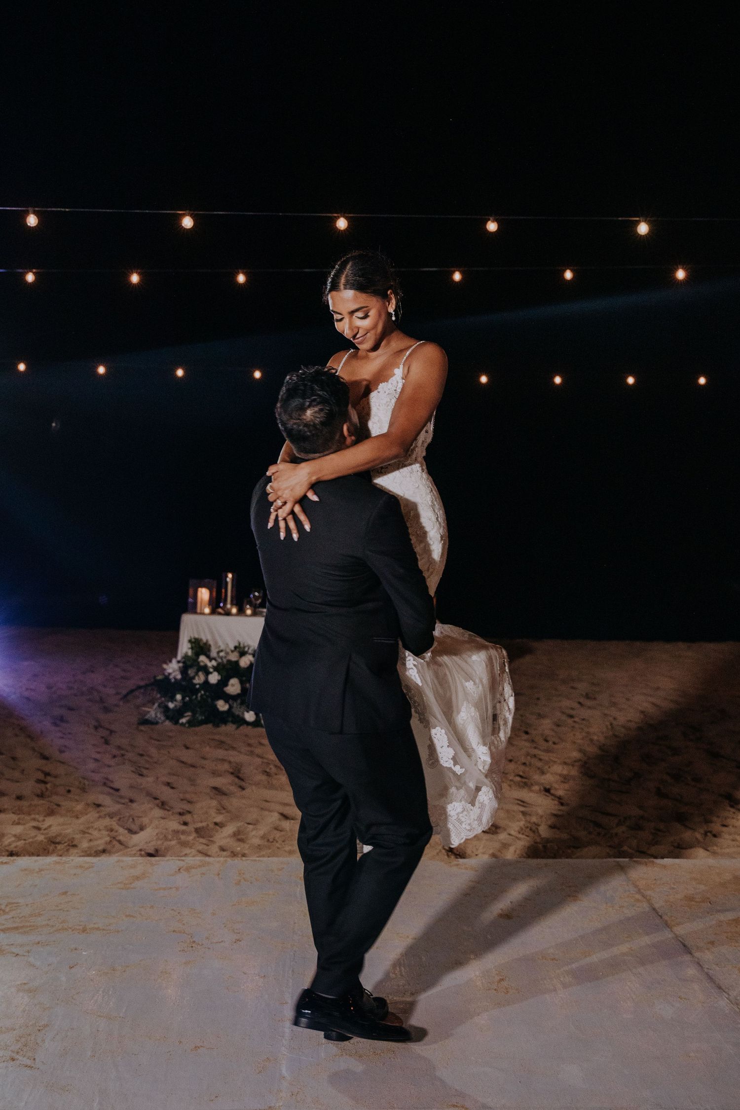 A couple shares a romantic dance moment on the beach at night under string lights.