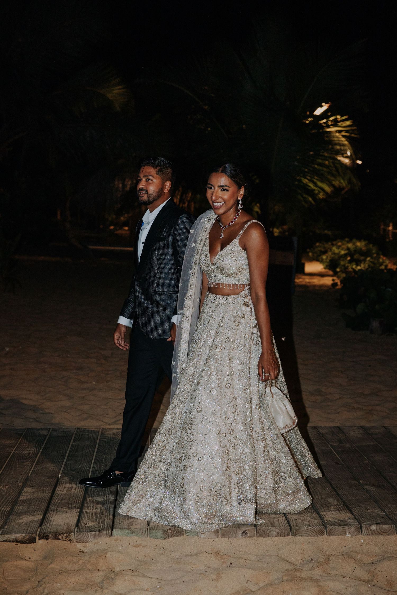 Couple in formal attire walking along a dimly lit beachfront pathway at night.