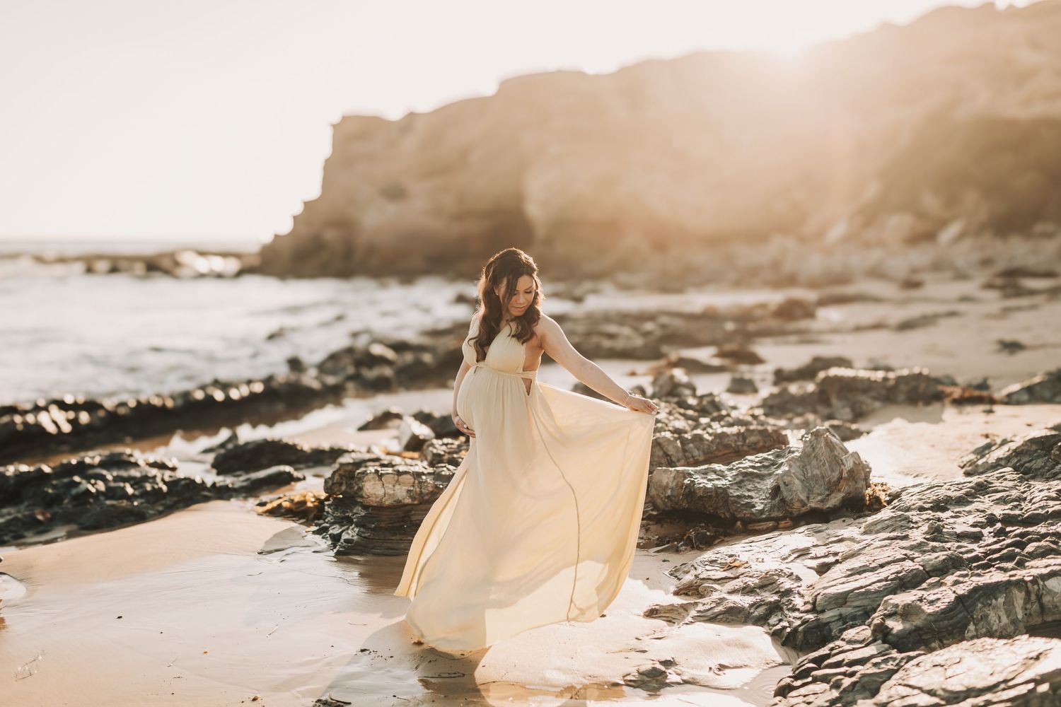 A flowing white dress catches the sunlight on a rocky beach at sunset with coastal cliffs in the background.