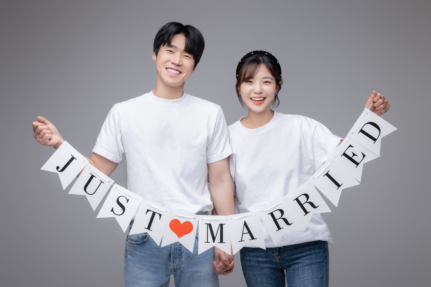 Two people in white t-shirts holding up a 'JUST MARRIED' banner together against a gray background.