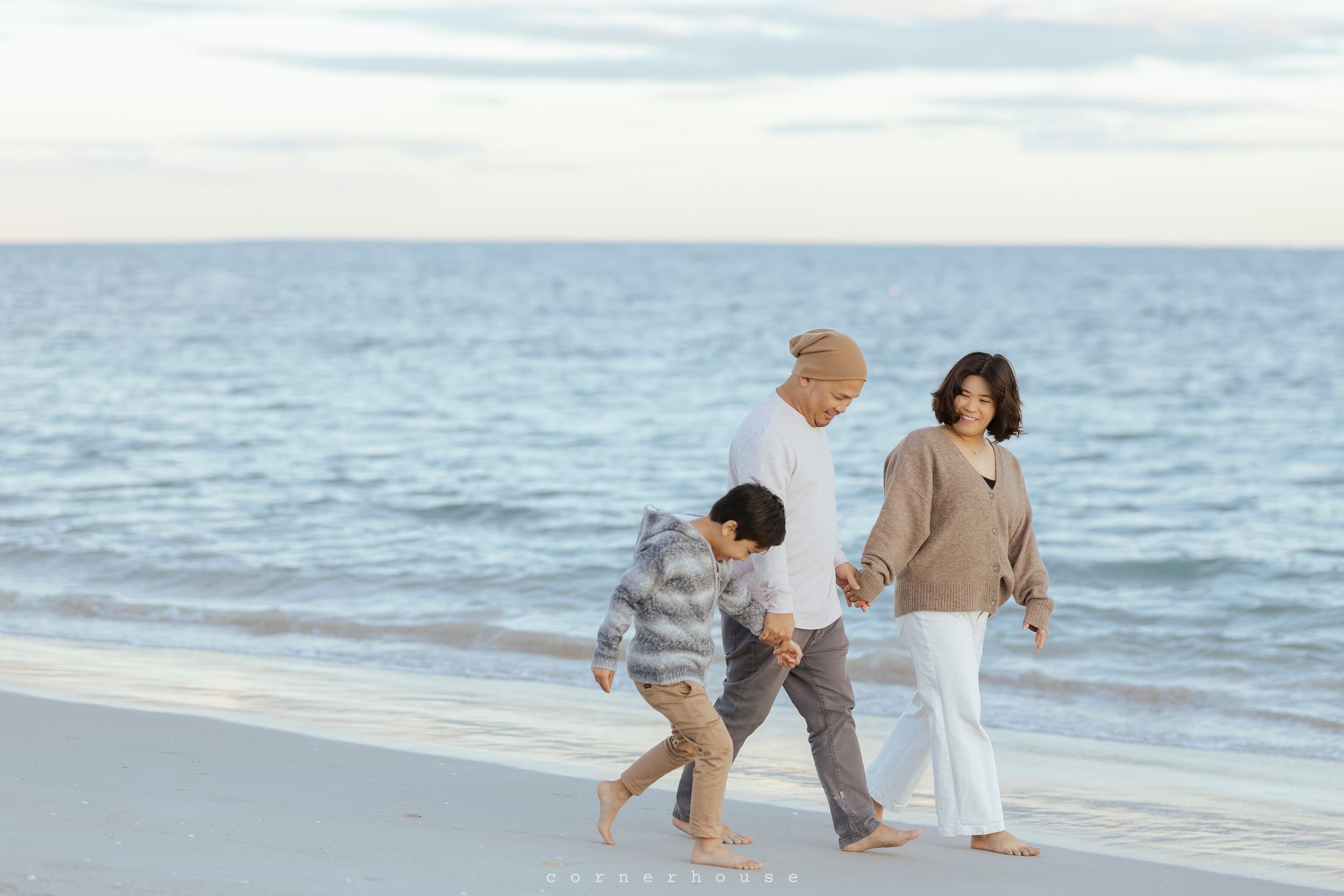 People in beige clothing walk along a scenic beach with waves and sand during sunset, creating silhouettes against the horizon.