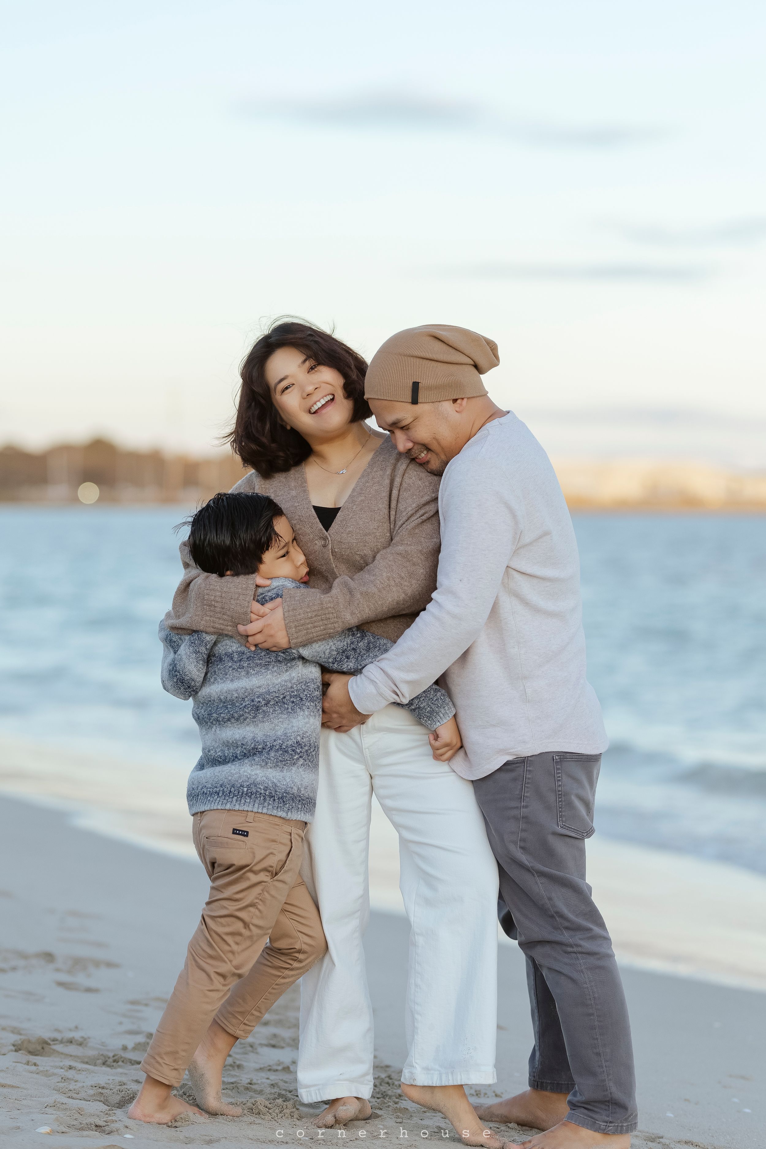 A family of three embraces while standing on a beach at sunset with ocean waves in the background.