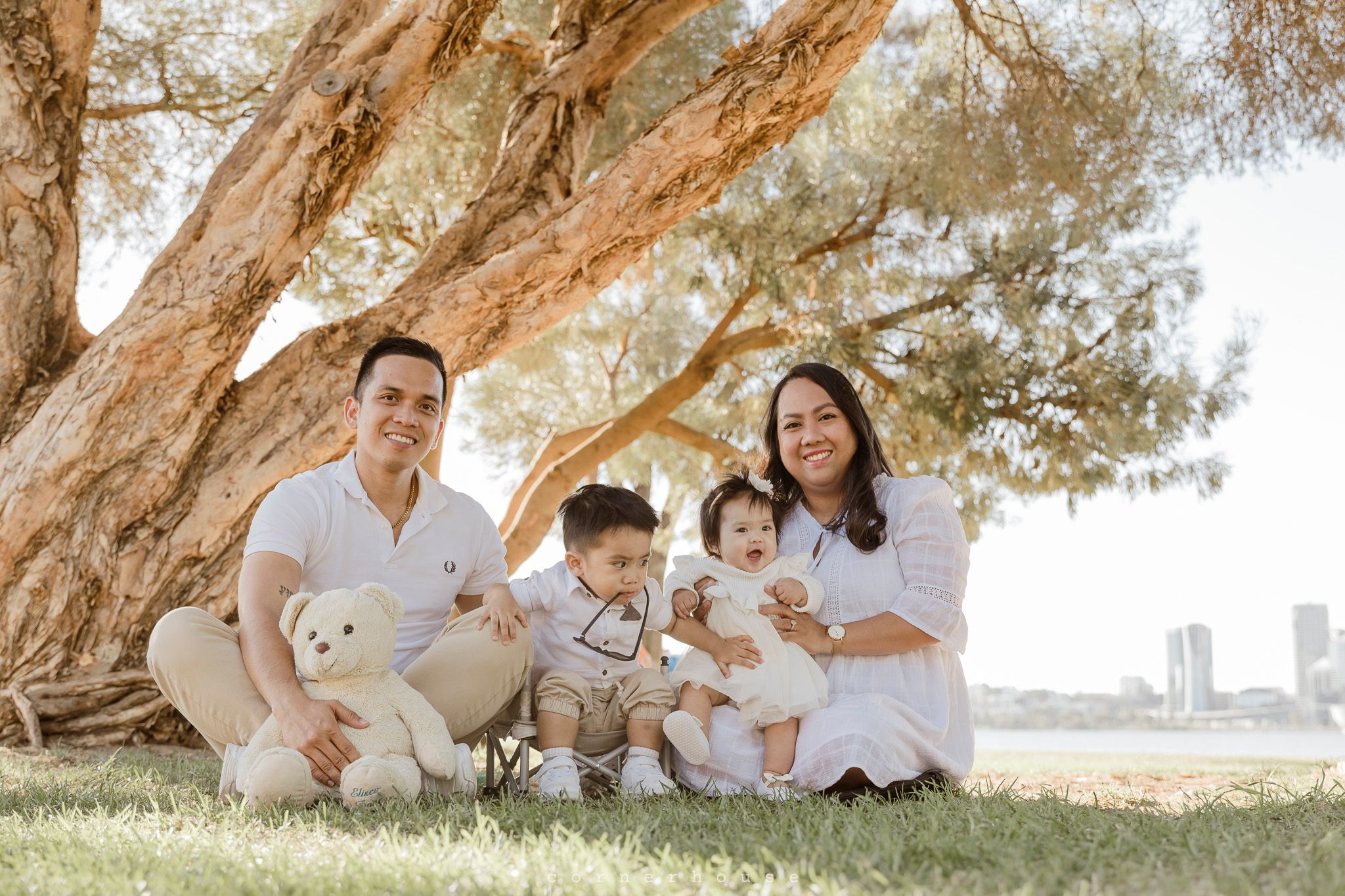 A family sitting together with their pets on grass under a large twisted tree trunk by the waterfront.