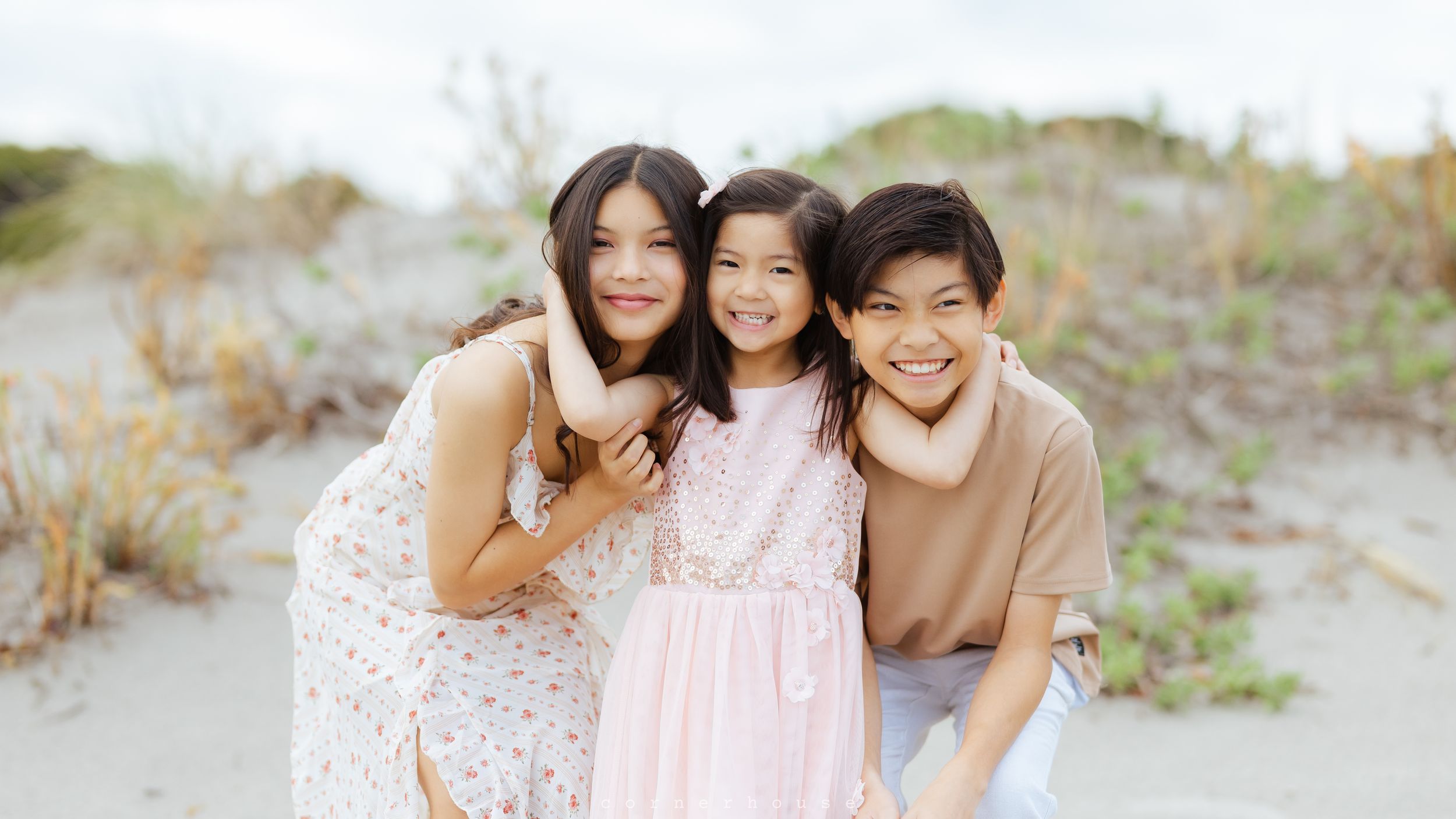 A group of three people in casual attire pose together outdoors on a sandy beach area with natural foliage.