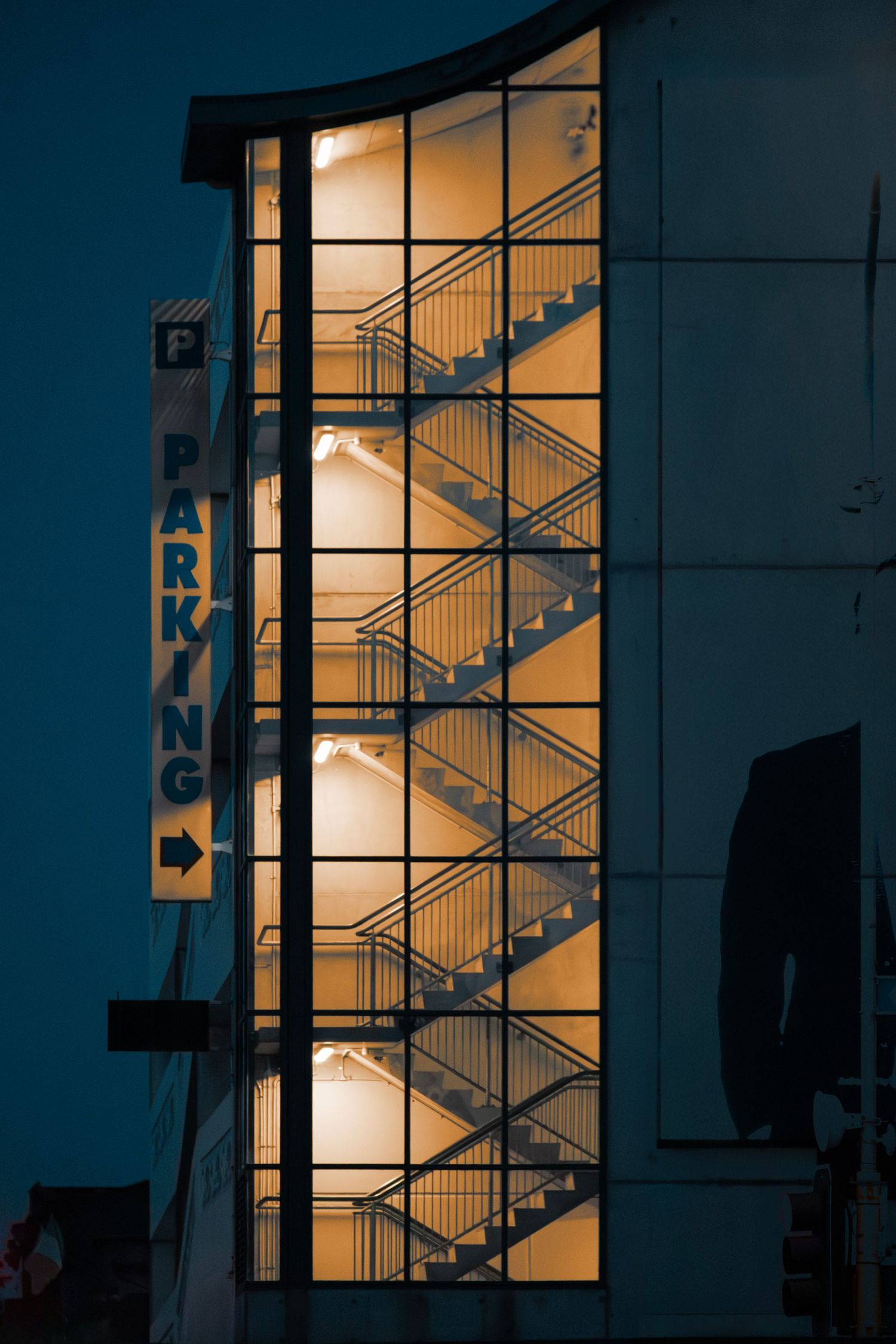 Glass-encased stairwell of a multi-storey parking structure, glowing with warm yellow light against the night sky.