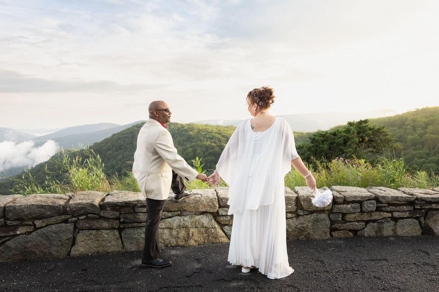Couple holding hands on a stone wall overlooking misty mountains during a scenic outdoor wedding ceremony.