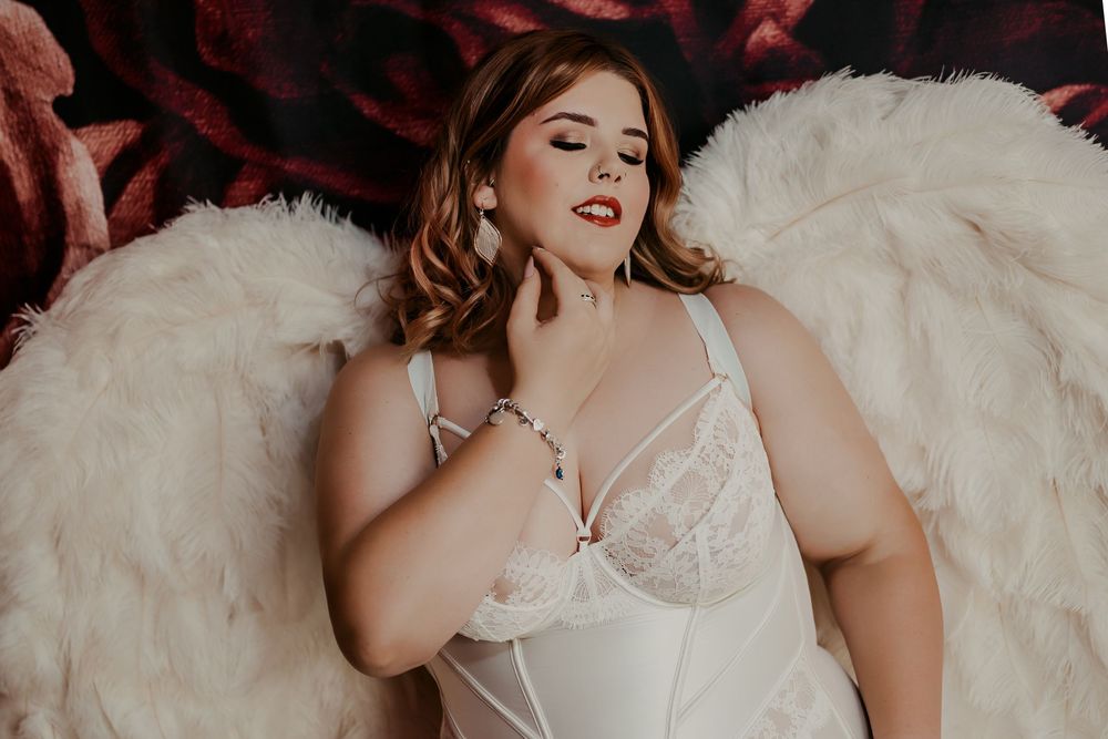 A figure in white lace lingerie poses with large feathered angel wings against a dark red background.