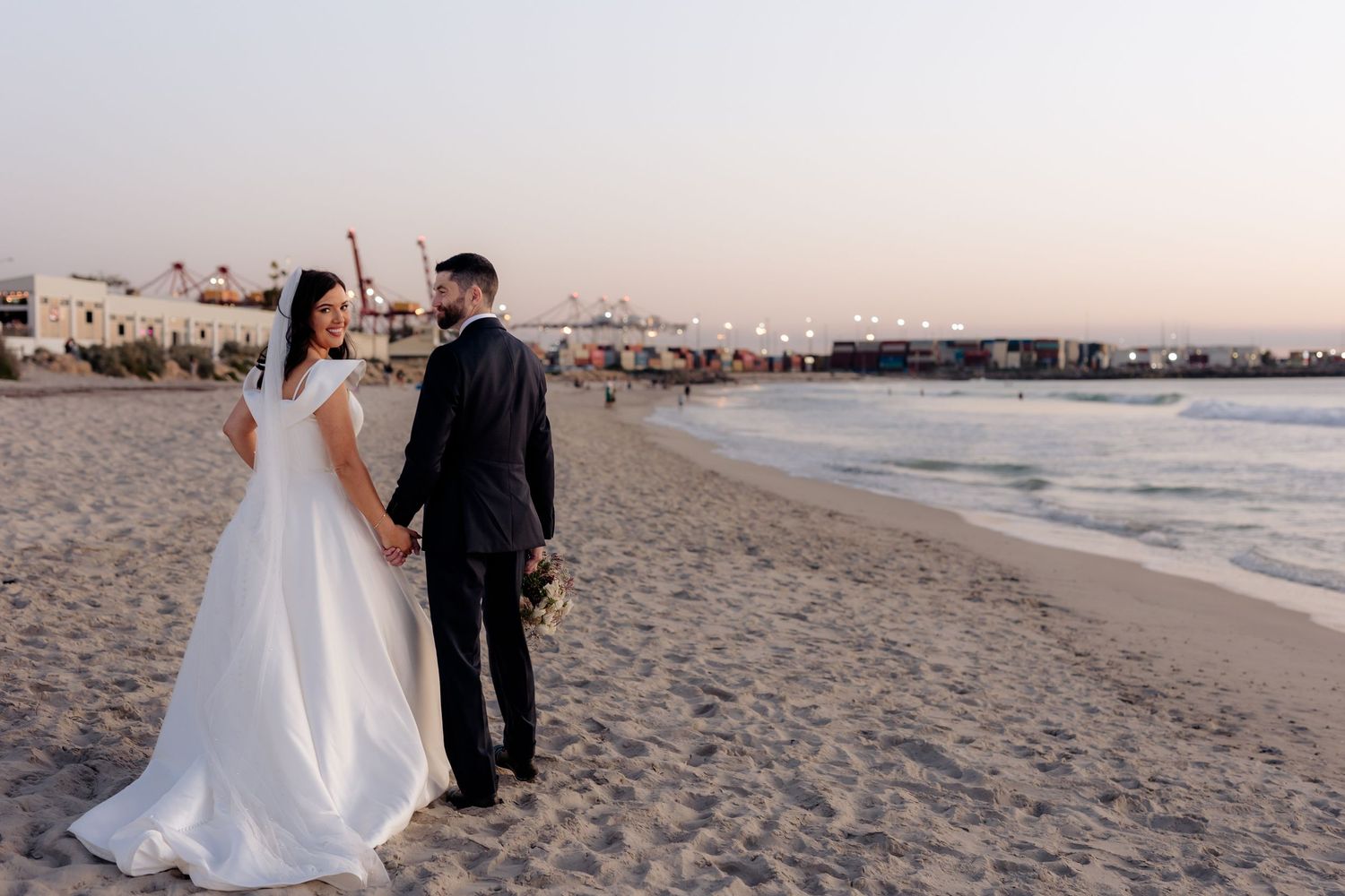 A newlywed couple shares romantic moments during sunset on a sandy beach with ocean waves in the background.