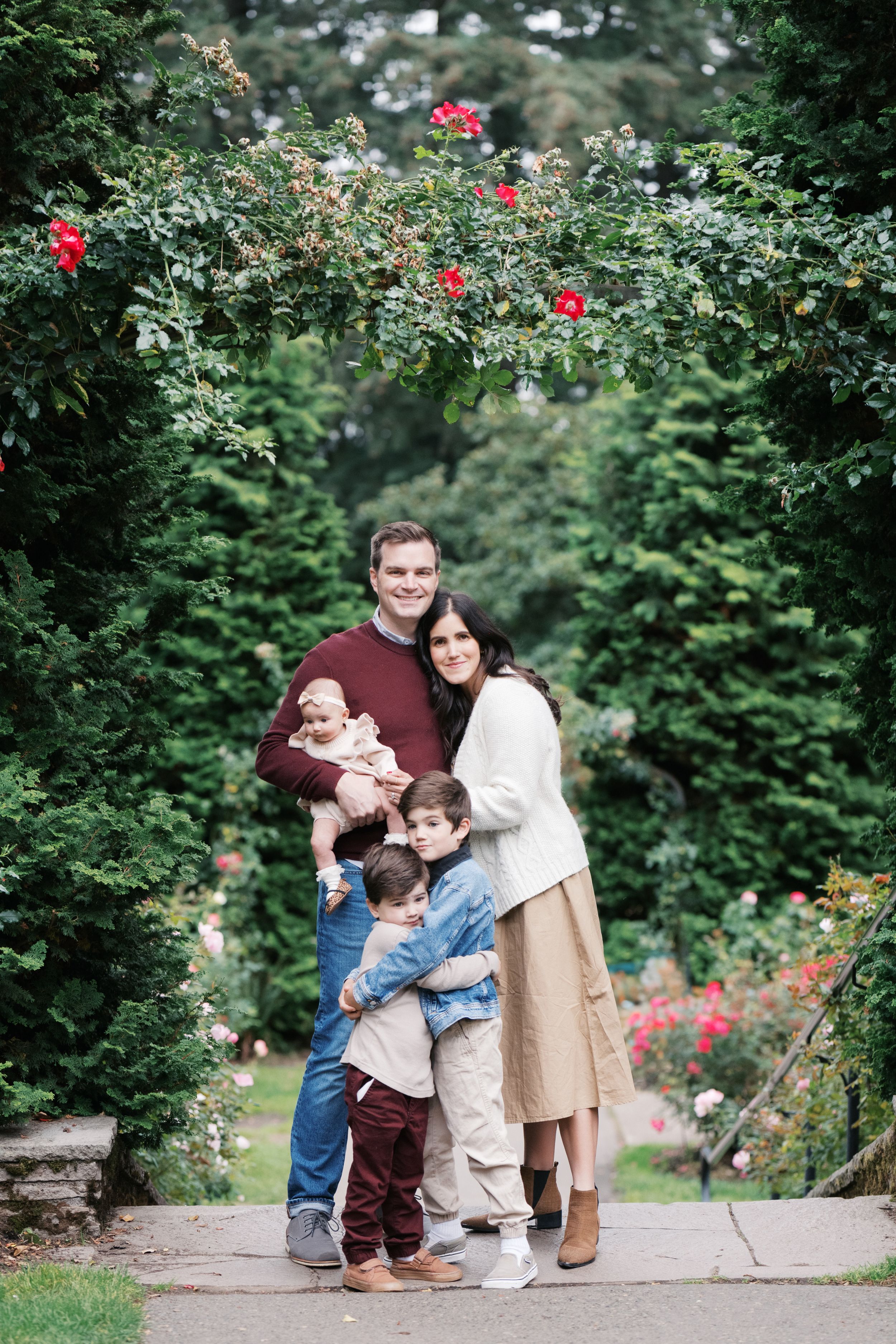 A family poses together under a rose archway in a garden setting on a stone path.