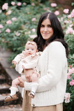 A baby in a cream outfit is held against a backdrop of pink roses and greenery.