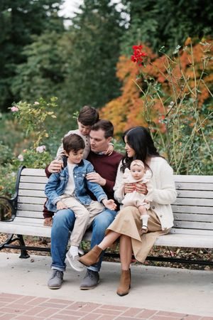 A family sits together on a wooden bench in a garden with red flowers and fall foliage in the background.
