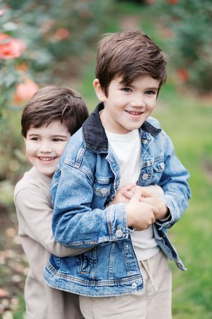 Two children in casual clothing share a playful moment together in front of flowering bushes.