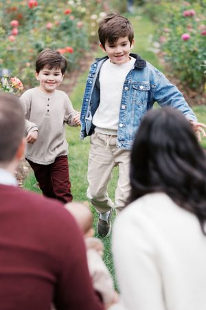 Two children playing and laughing in a rose garden while adults watch nearby.
