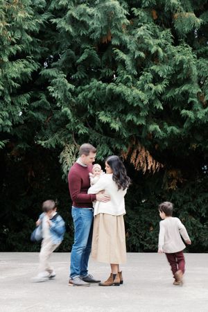 A family shares a tender moment by evergreen trees as children play in motion.