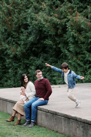A joyful family moment captured outdoors as a child jumps energetically while others rest on a concrete ledge.