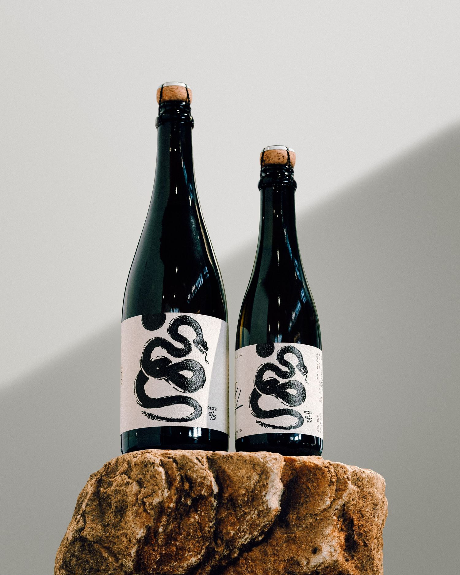 Two dark glass bottles with snake logo designs sit on a textured stone surface in dramatic lighting.