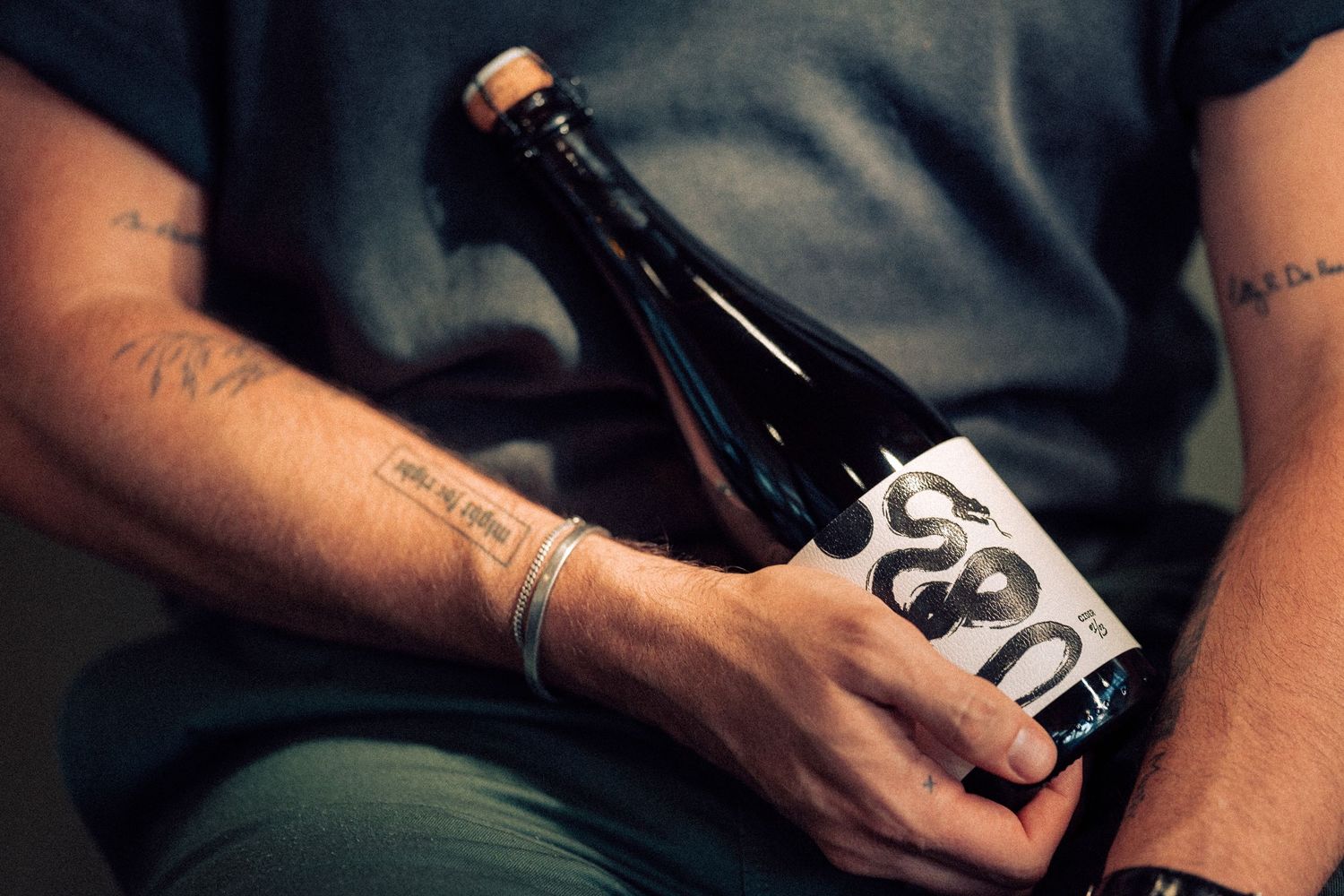 Tattooed hands hold a dark glass bottle with simple black and white minimalist label design against dark clothing.