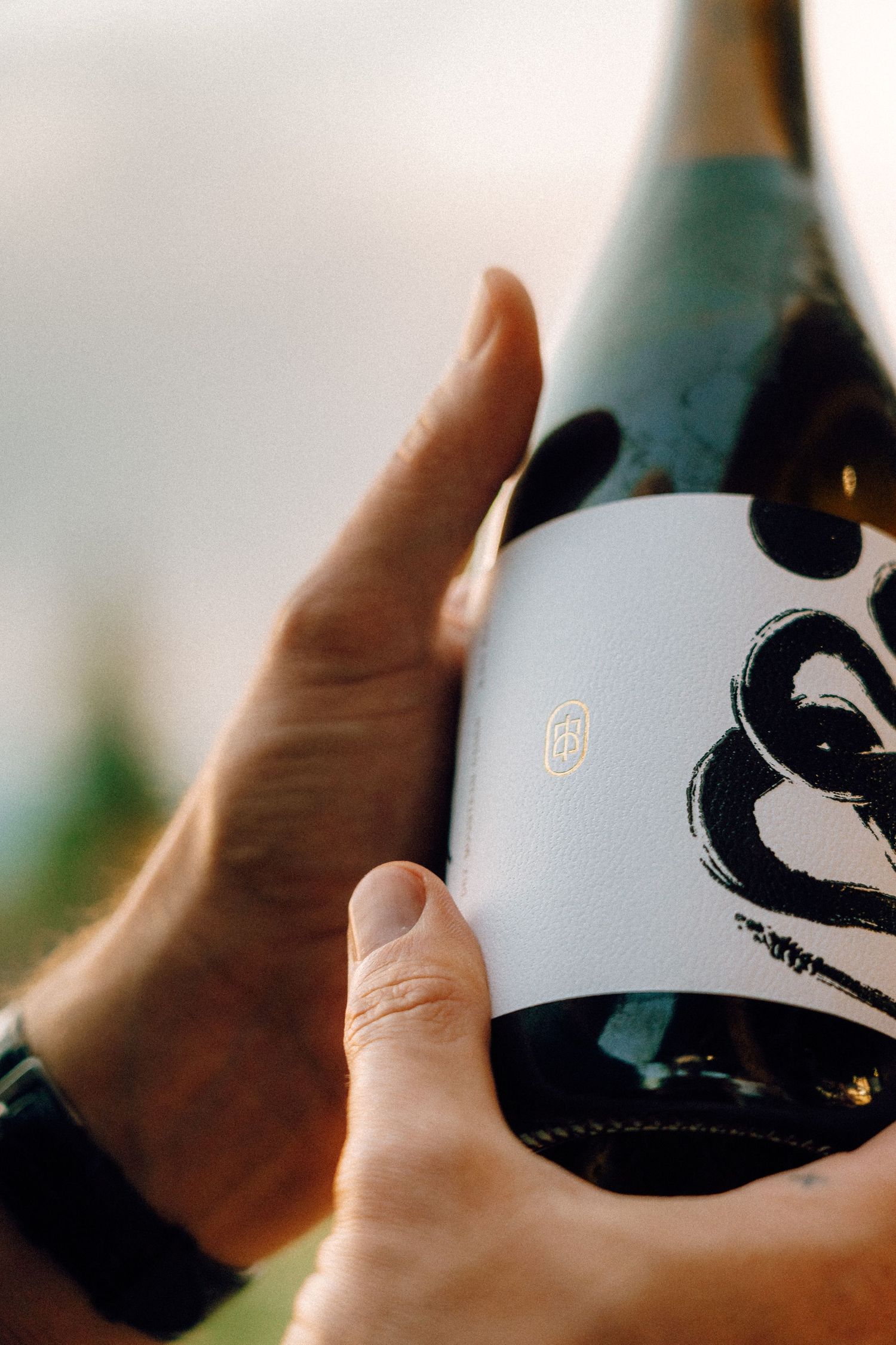 A glass wine bottle with black and white branding being held up against a blurred background.