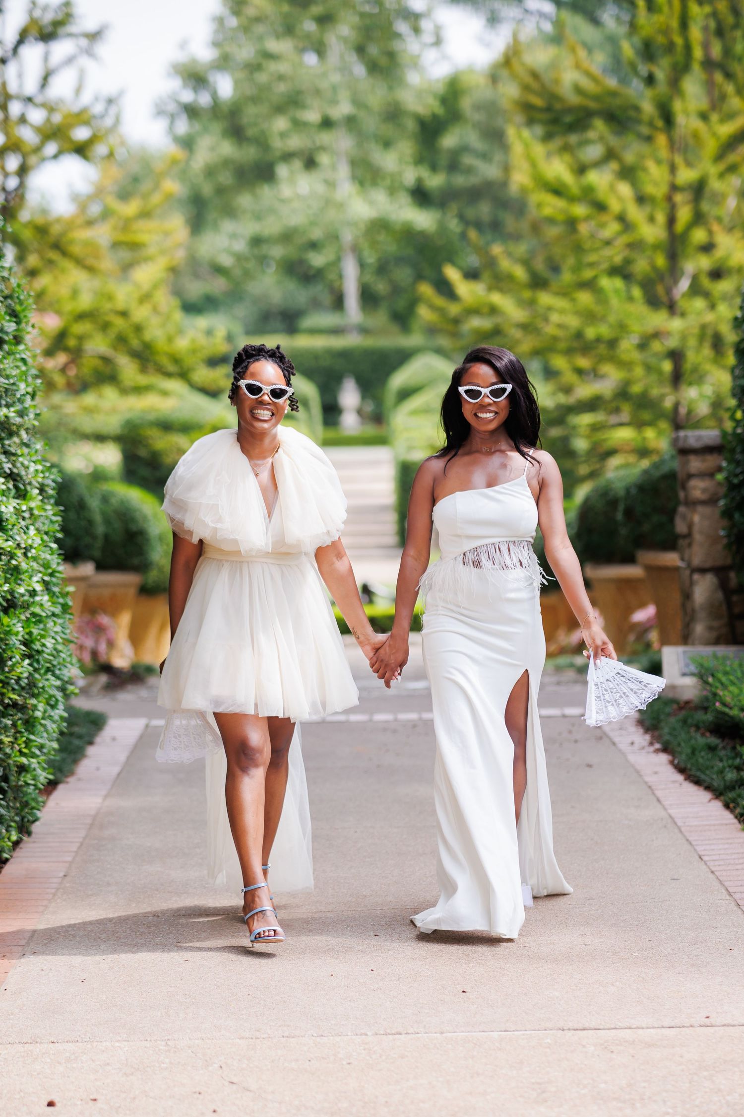 Two brides in white dresses walking hand in hand along a garden path during their elopement at the Dallas Arboretum
