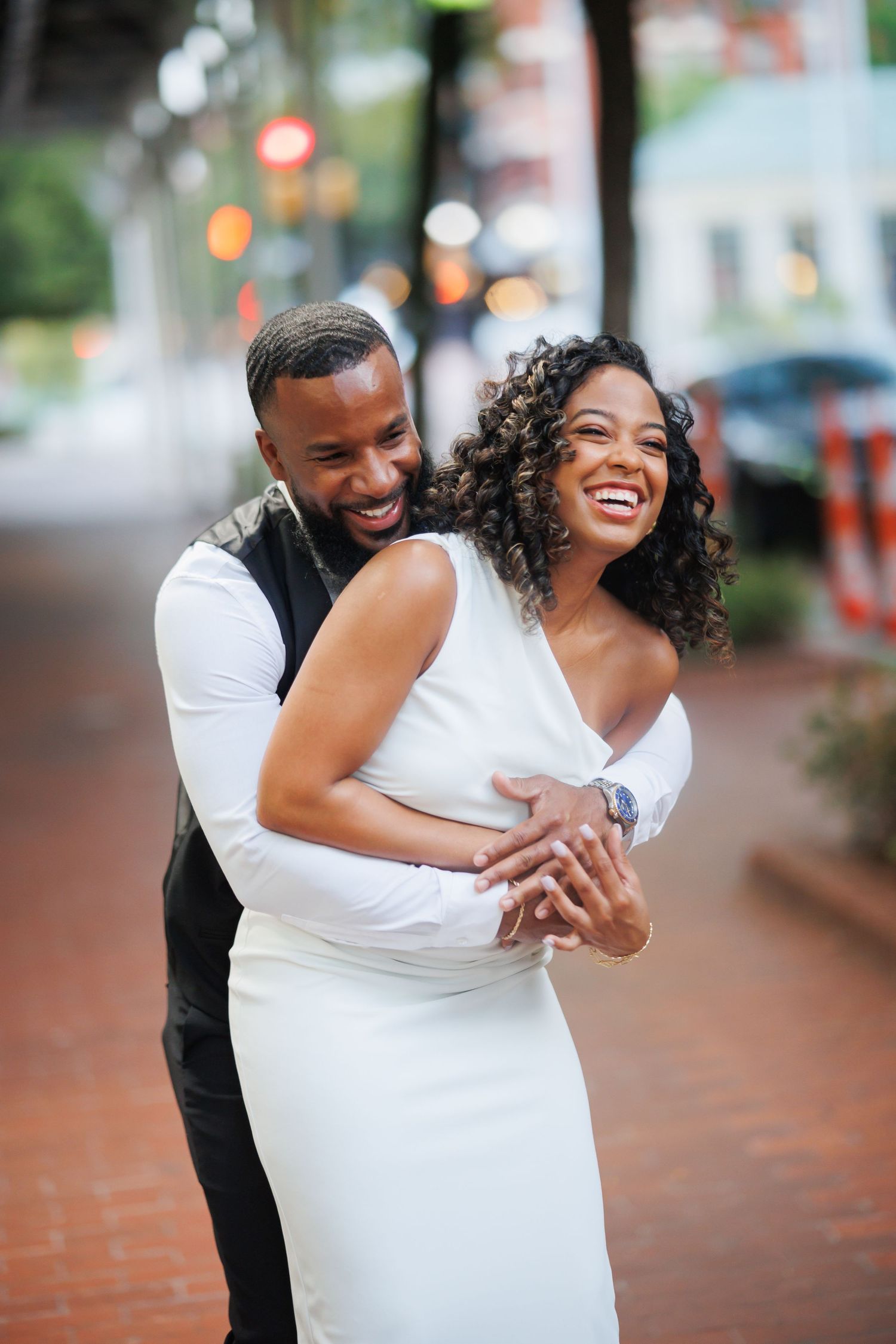 Happy couple shares a candid laughing moment together on a brick sidewalk in formal wedding attire.