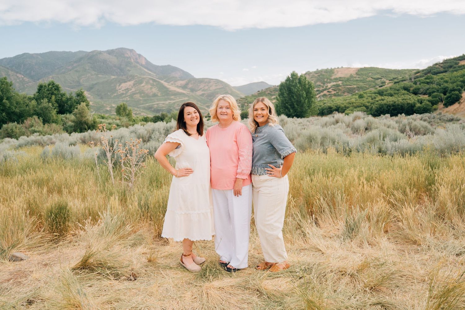 Three people in coordinating outfits stand together in a golden field with mountains in the background.