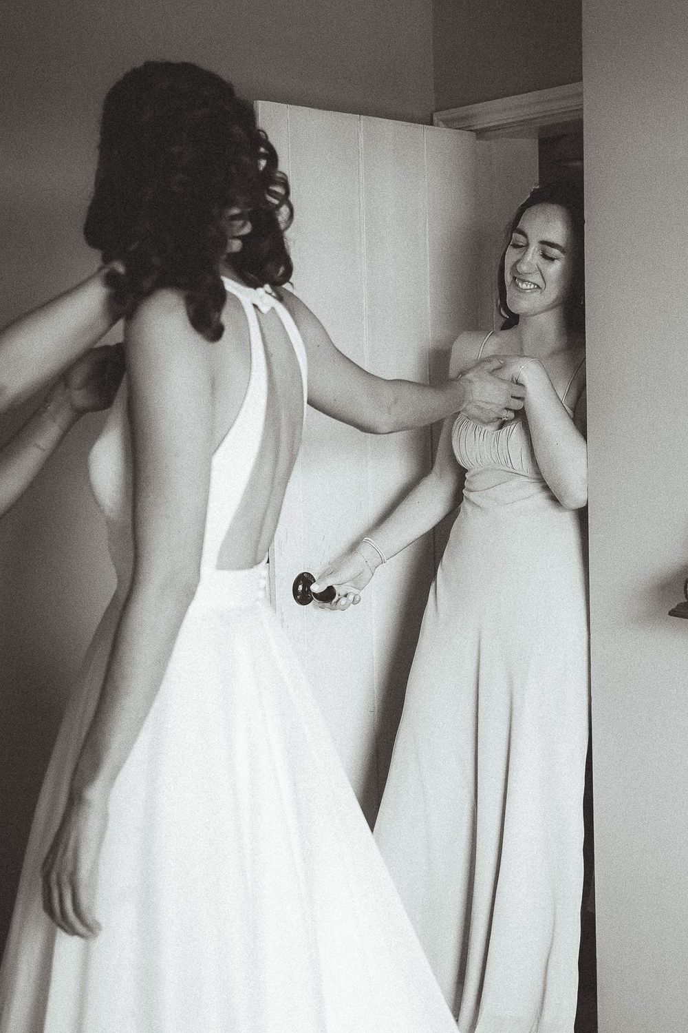 Two women in elegant white dresses share a joyful candid moment in black and white photography.