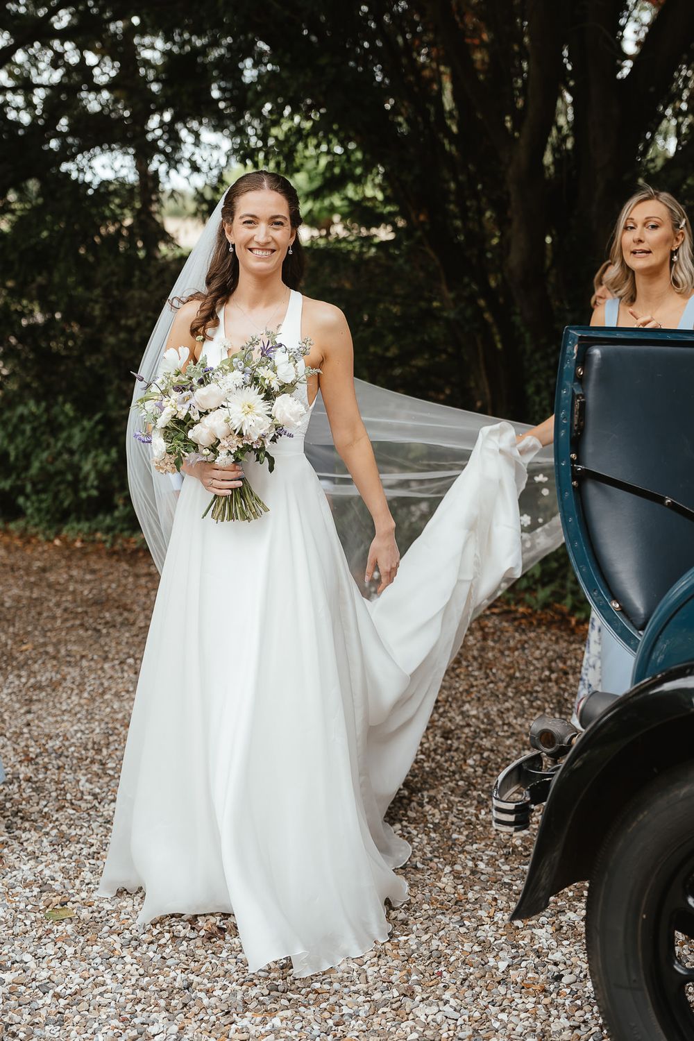 A bride in a flowing white gown holds a pastel bouquet while standing next to a vintage car on a gravel path.