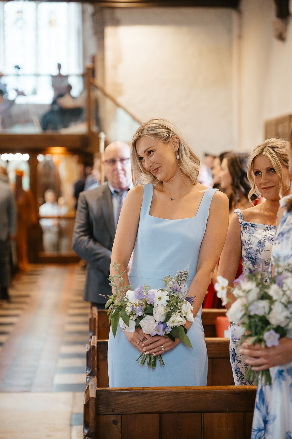 Wedding party members in pastel blue and floral dresses hold delicate bouquets during church ceremony.