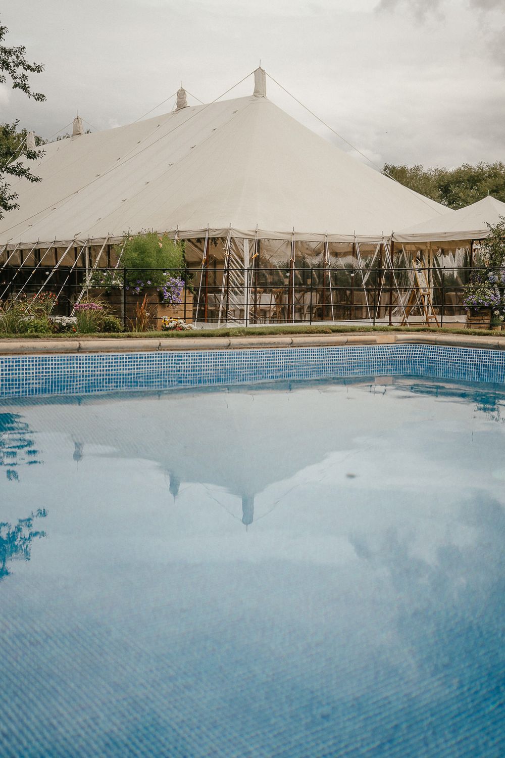 Large white wedding tent with clear sides overlooks a sparkling blue swimming pool at an outdoor venue.
