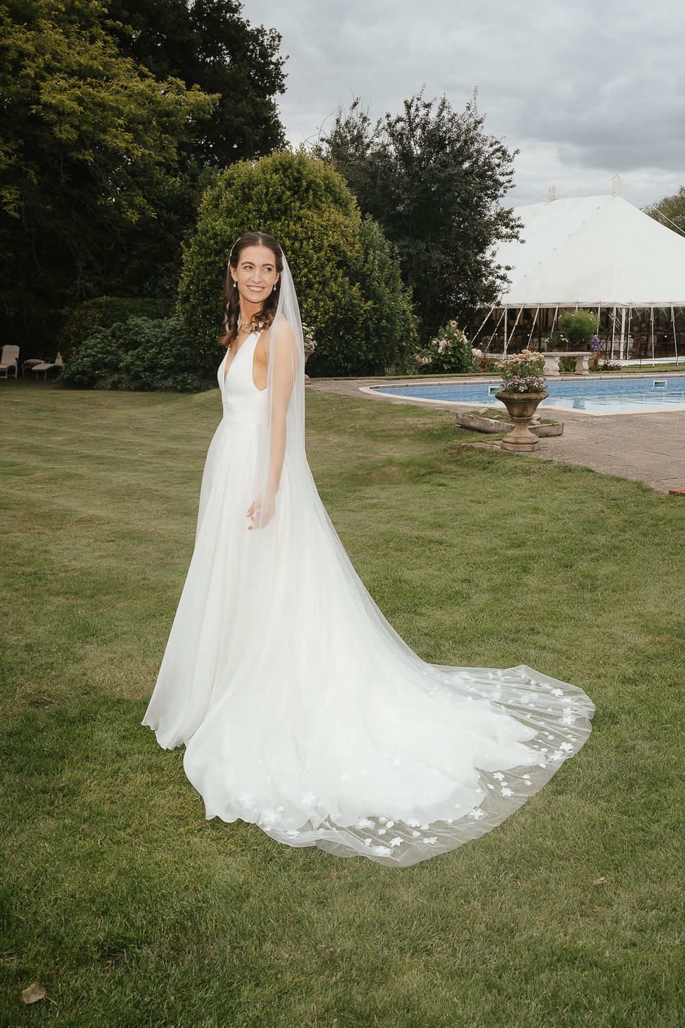 A white wedding gown with a flowing train on a manicured lawn with a tent and pool in the background.