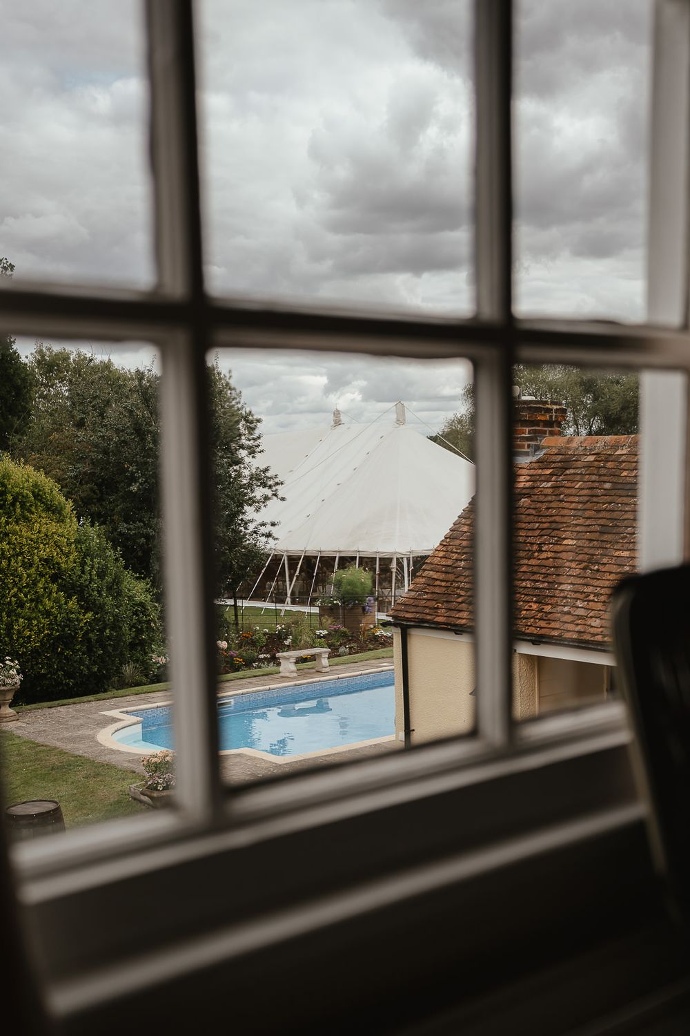 View through window panes of a backyard swimming pool and white event tent on a cloudy day.
