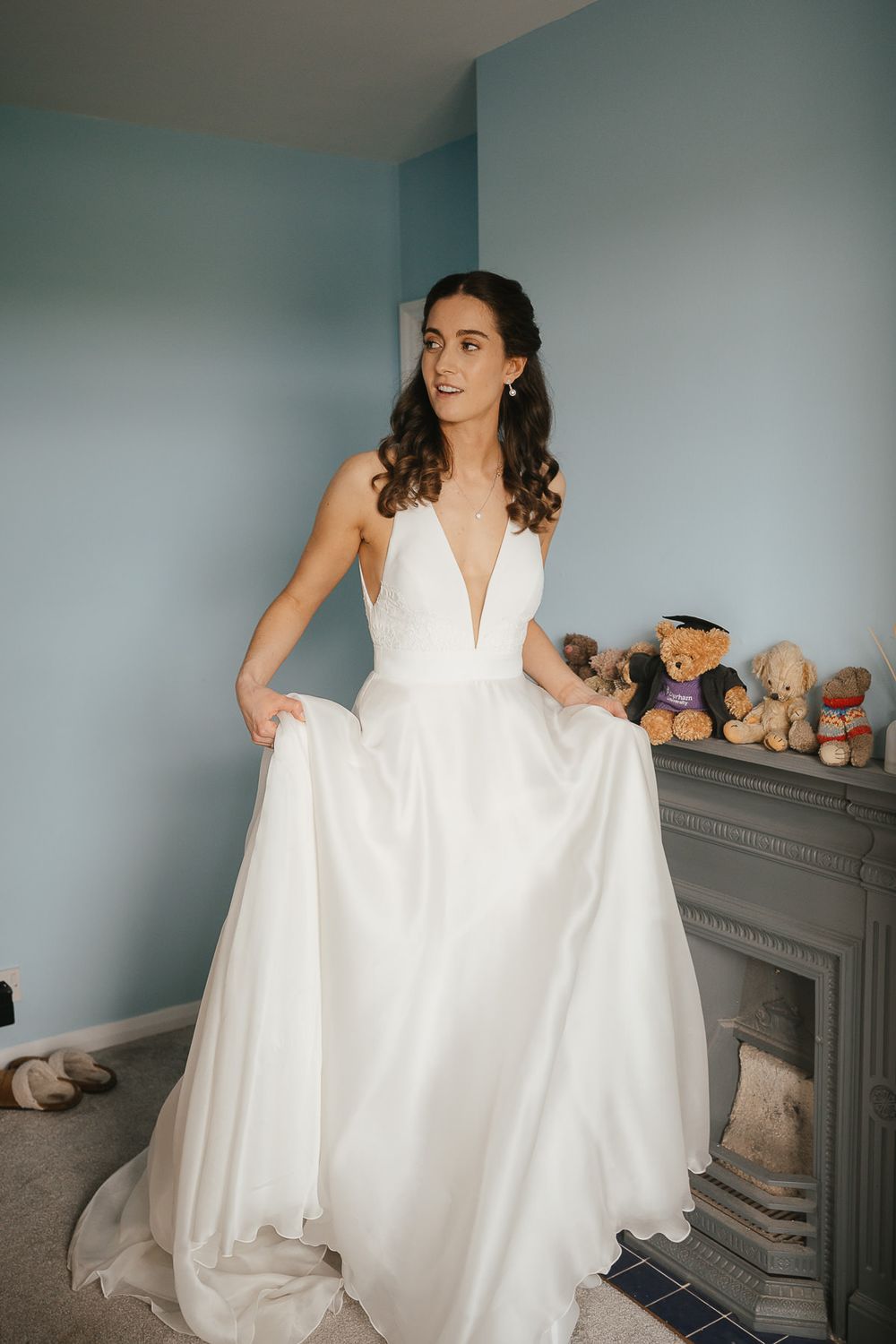 An elegant white wedding gown with plunging neckline and flowing skirt shown in a bedroom with stuffed animals on fireplace mantle.