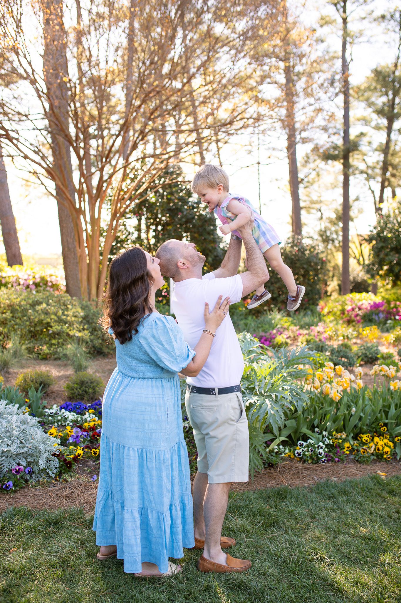 A family plays together on a garden path surrounded by tall pine trees and flowering plants at WRAL Azalea Garden