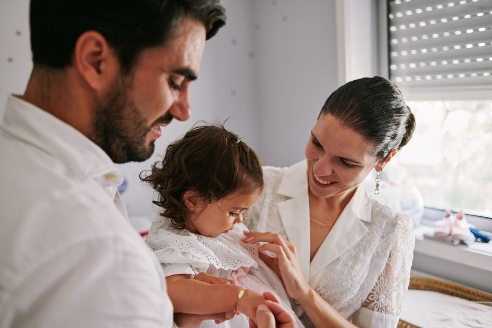 Smiling parents in white attire attending to their young daughter during a baptism celebration.