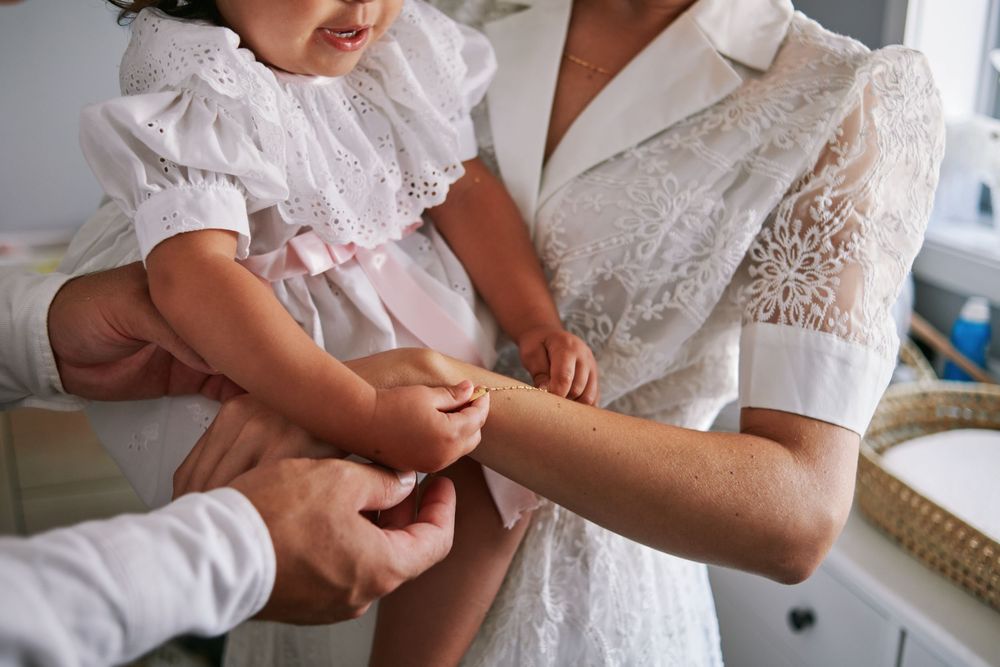 Woman in white lace dress holding a toddler girl in pink dress during a baptism preparation ceremony.