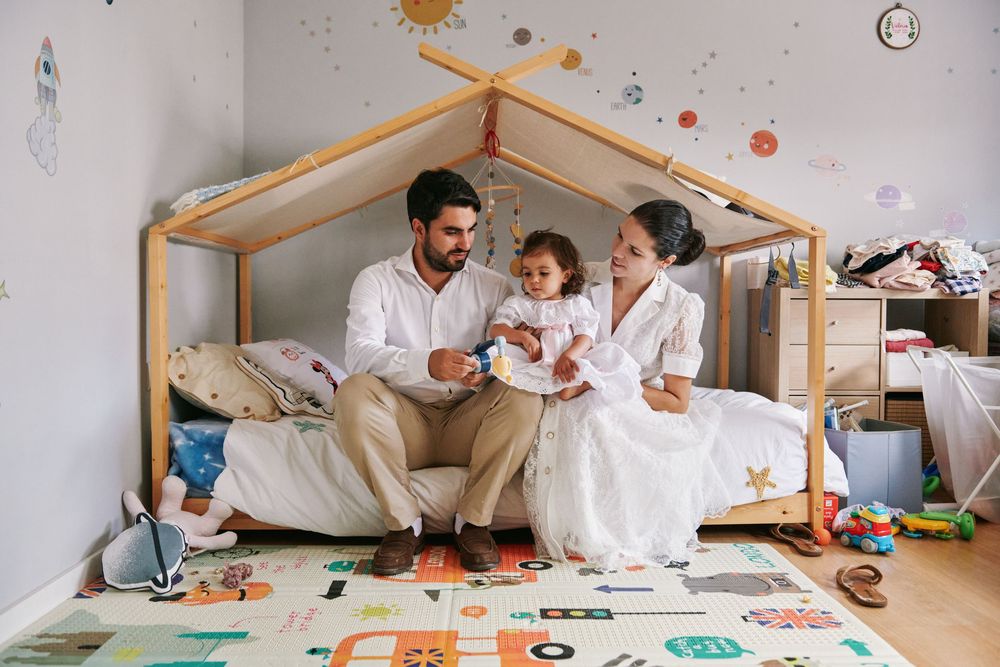 Happy young family with a toddler sits together on a wooden house-frame bed in a decorated nursery.