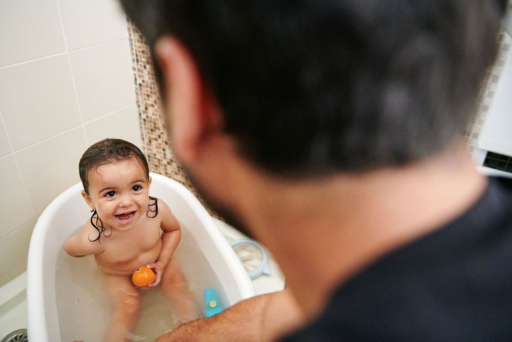Smiling baby enjoys bath time in a small tub while father watches lovingly from the foreground.