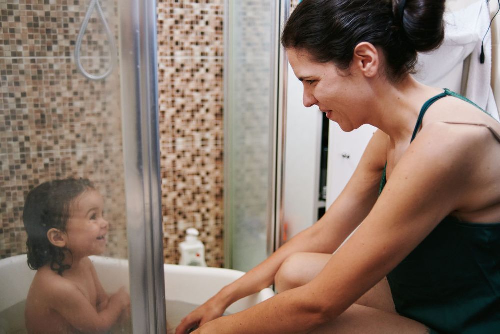 Mother in green top leaning over bathtub smiling at her young toddler during bath time.