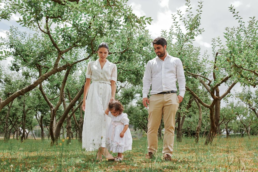 Family of three walking together in a lush green orchard, parents dressed in white and beige.