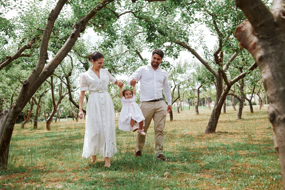 Happy family with mother in white dress and father swinging their toddler in a green orchard.