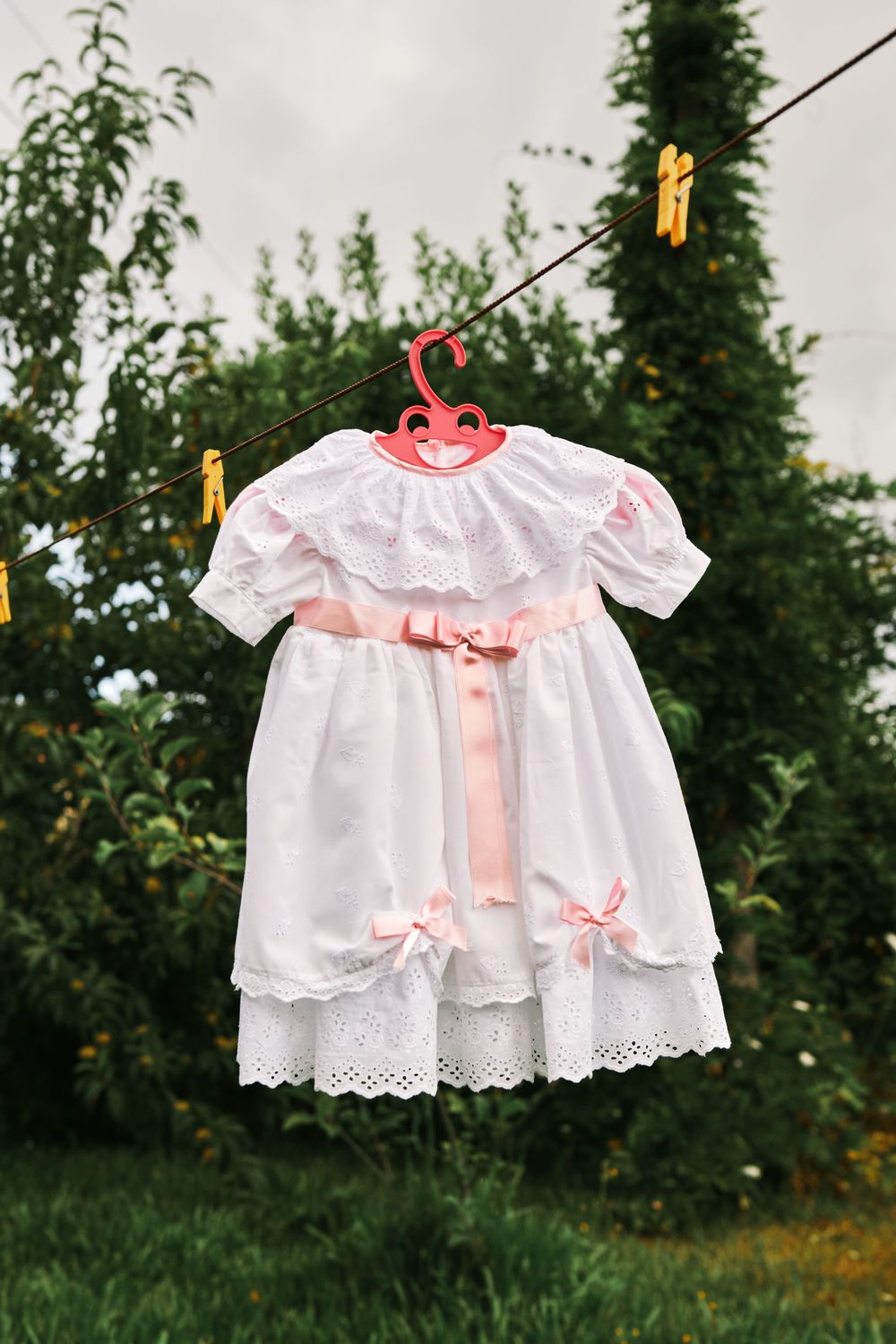 Delicate white baby girl dress with pink ribbons and bows hanging on a clothesline outdoors.