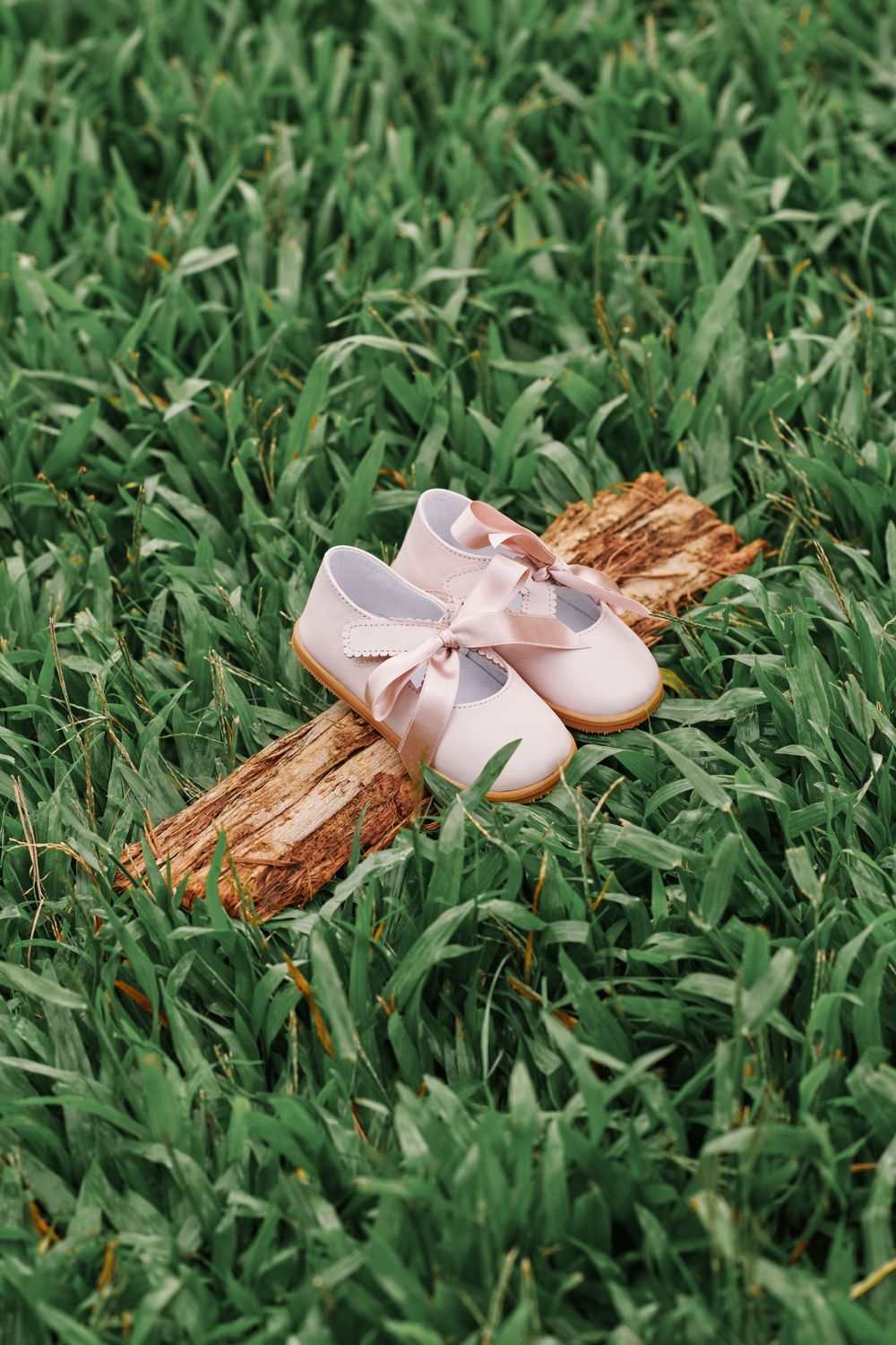 Tiny pink baby ballet shoes with satin bow rest on a wooden cross in green grass.