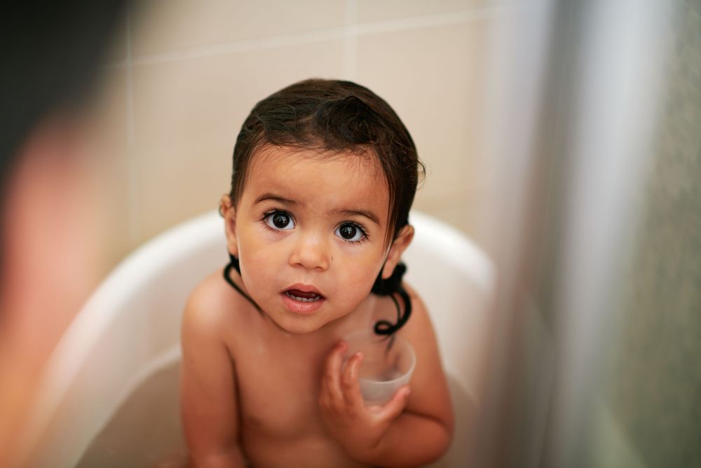 Adorable baby with dark eyes sitting in a white bathtub during bath time.