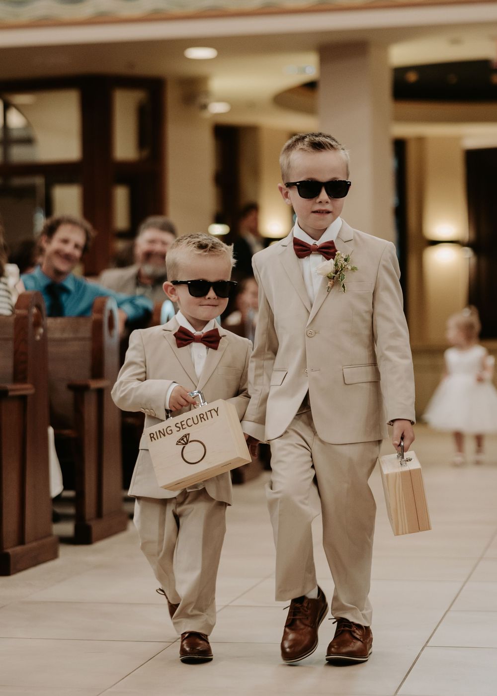 Two young ring bearers in tan suits and sunglasses walking down a hallway.